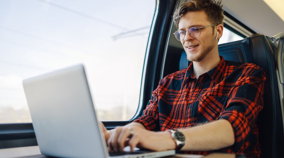 Tourismusprofi Porträt eines jungen Passagiers, der in der Nähe des Zugfensters sitzt und auf einem Laptop tippt, während er ihn lächelt und mit dem Zug fährt. Ein glücklicher Hipsterreisender arbeitet auf seiner Bahnreise an einem Laptop.