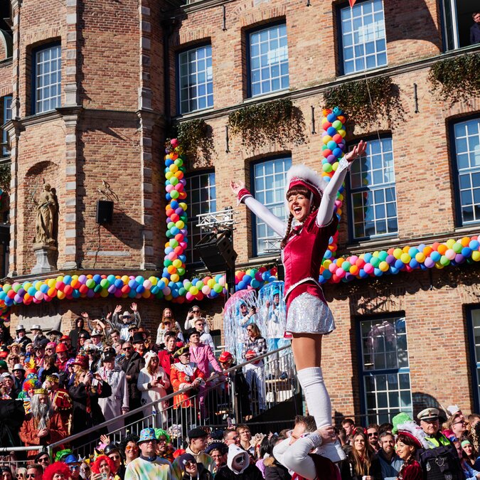 © Visit Düsseldorf - Markus Luigs Fröhliche Karnevalsfeier vor einem historischen Backsteingebäude in Düsseldorf: Eine kostümierte Person hebt jubelnd die Arme, während sie auf den Schultern einer anderen steht. Bunte Luftballons schmücken die Fassade, und zahlreiche verkleidete Menschen feiern auf dem Platz davor. | © Visit Düsseldorf - Markus Luigs