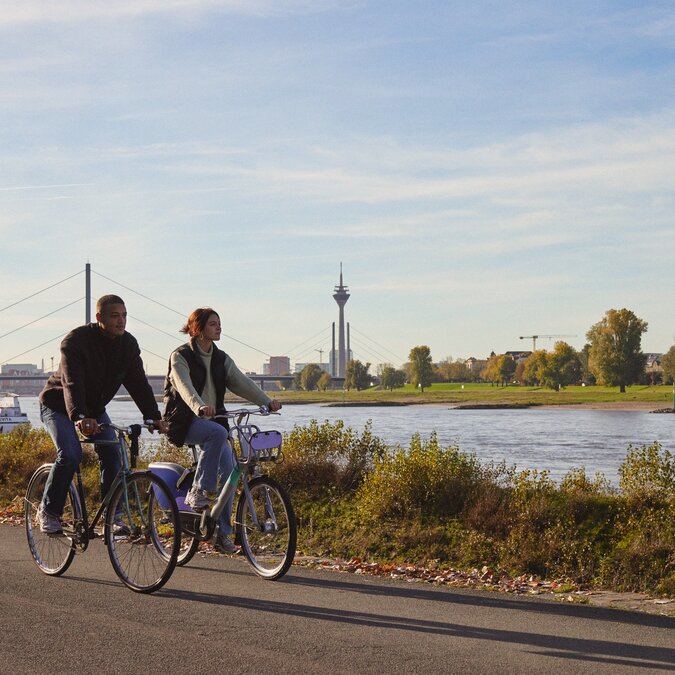 Fahrradtour am Rhein | © Düsseldorf Tourismus – React-EU Zwei Personen radeln am Rheinufer mit Blick auf den Rheinturm. | © Düsseldorf Tourismus – React-EU