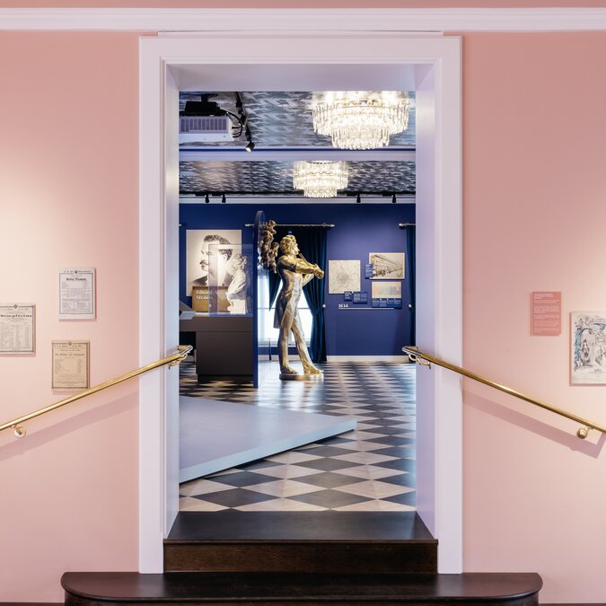Ausstellung im Johann Strauss Museum | © Wien Tourismus |Gregor Hofbauer Blick durch einen rosafarbenen Eingangsbereich in einen Ausstellungsraum mit Skulpturen und historischen Exponaten. | © Wien Tourismus |Gregor Hofbauer