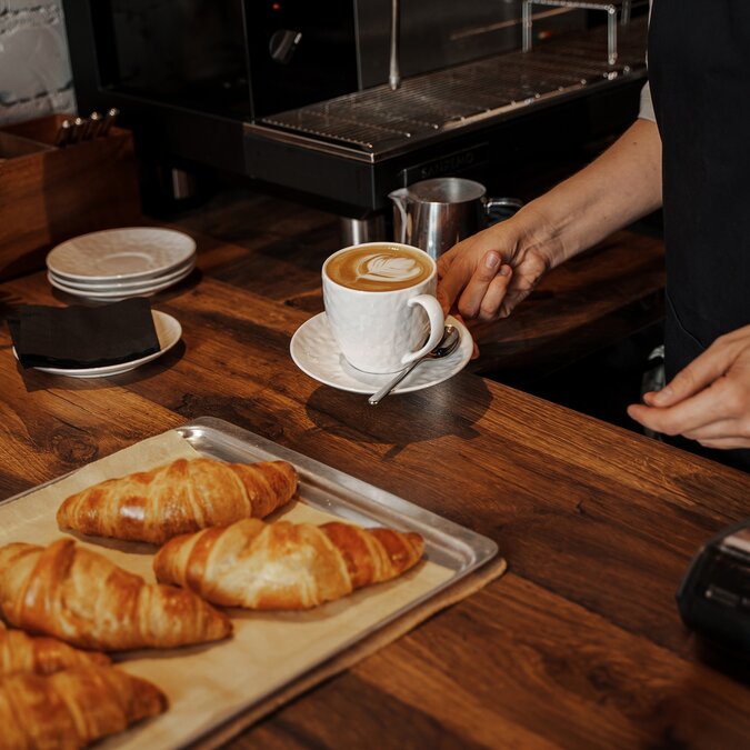 Café Hygge | © Shutterstock 2393949781 Ein Barista platziert einen Cappuccino mit Latte Art neben frisch gebackenen Croissants auf einer Holztheke in einem gemütlichen Café-Ambiente | © Shutterstock 2393949781