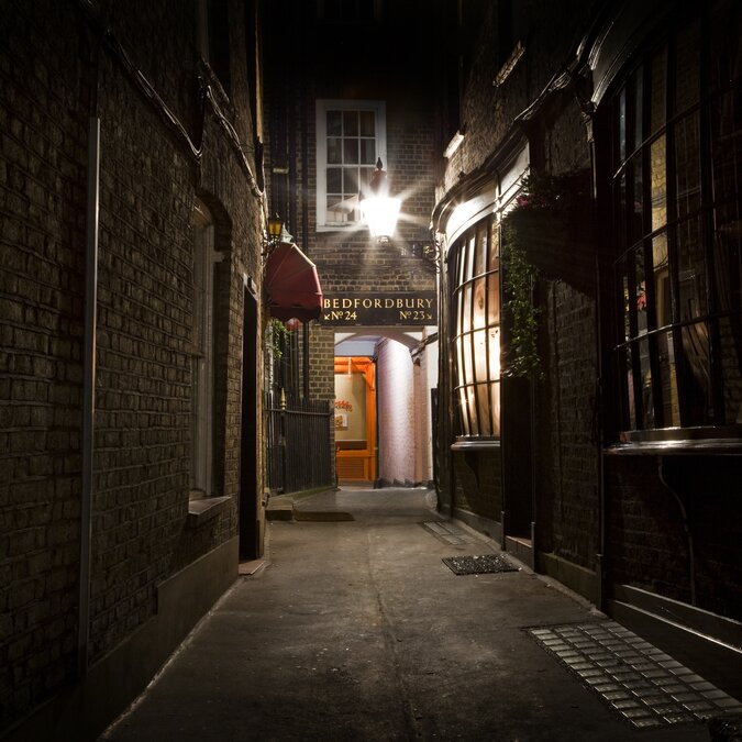 Stimmungsvolle Nachtaufnahme einer schmalen Gasse in London. | © shutterstock // 125129336 Eine enge, dunkle Gasse mit Backsteinmauern und einem beleuchteten Eingangsschild mit der Aufschrift "Bedfordbury". | © shutterstock // 125129336